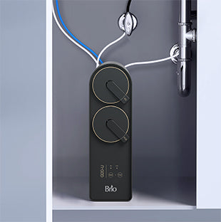 A black Brio water filtration system is installed under a sink, connected to pipes and water lines, with two circular dials and LED indicators on its front panel.